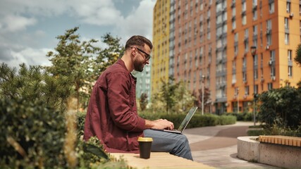 Happy and young. Side view of handsome man with stubble in casual clothes and eyeglasses working on laptop while sitting on the bench outdoors