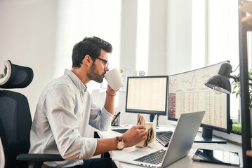 Coffee break. Young bearded businessman or trader is drinking a coffee and eating a sandwich while looking at monitor screen with financial data in his modern office - obrazy, fototapety, plakaty