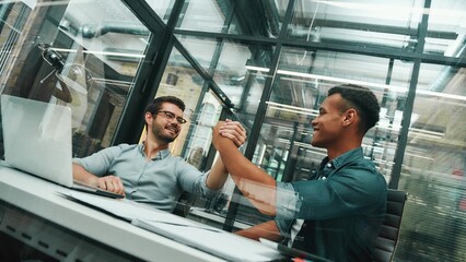 Glad to work with you Two young and handsome men shaking hands and smiling while working in modern office