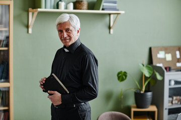 Waist up portrait of smiling senior priest looking at camera and holding Bible while standing in...
