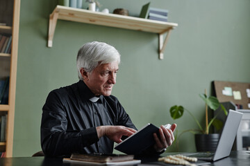 Side view portrait of white haired senior priest holding Bible while giving online religious support talk via video chat, copy space - obrazy, fototapety, plakaty