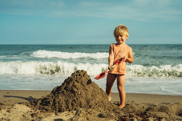 beautiful little boy 3 years old blond digs sand at the sea with a shovel