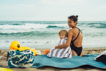 mom and son are relaxing on the beach sitting in front of the sea on a mat on a sunny summer day