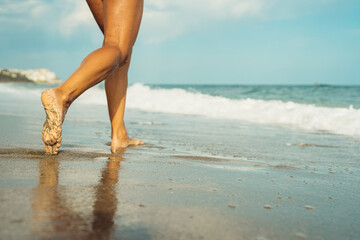 Fototapeta na wymiar beautiful tanned female legs walk on the sand against the backdrop of the sea on the beach on a sunny day