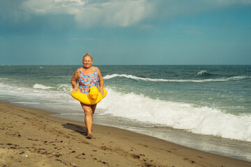 beautiful grandmother 60 plus blonde with a smile on the beach with a lifebuoy yellow duck walks on the sand