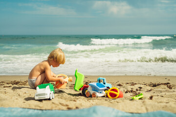 boy 3 years old blond plays on the seashore in the sand with plastic toys against the background of the sea