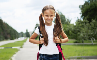 Portrait of happy girl with school bag standing outdoor. Little happy school girl going to school, back to school, education concept. Cute female student smiling and looking at camera