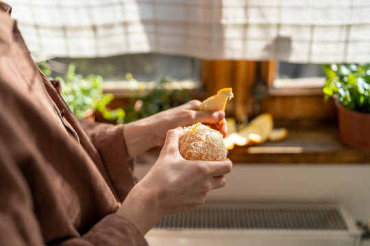 Girl feel good mood peels ripe tangerine puts orange on wooden windowsill with green plants. Woman enjoys fresh juicy citrus aromas from fruit while standing in cozy apartment. Closeup