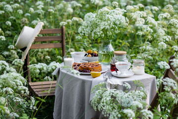 Tea time with sweet homemade cherry cake and herbal beverage in summer garden outdoors. Table covered with linen tablecloth, white porcelain cups. English style
