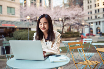 Young happy Asian business woman professional or student sitting at street table using laptop hybrid working or remote learning online typing on computer technology in city park.