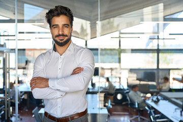 Confident young Latin business man standing in office arms crossed, portrait. Smiling Hispanic...
