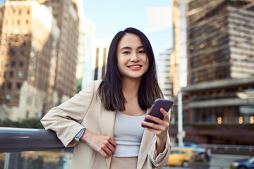 Young cute happy pretty Asian business woman professional manager leader with cellphone, smiling employee standing on big city street holding mobile phone, using smartphone, portrait.
