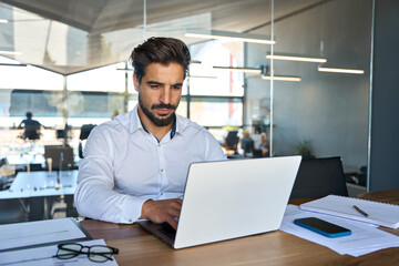Busy young Latin business man looking at laptop at work. Focused young businessman company executive manager investor using computer sitting in office thinking of investment plan checking digital data - obrazy, fototapety, plakaty