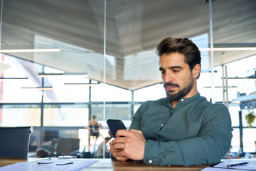 Fototapeta na wymiar Busy young Latin business man using mobile cell phone at work in office. Serious male executive businessman manager sitting at desk holding smartphone working on cellphone at workplace.