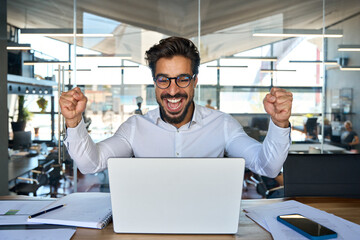 Excited professional young Latin business man executive happy about financial market growth or work results, corporate goals achievement getting email with good news sitting with laptop in office.