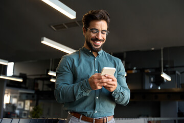 Smiling handsome Latin business man executive or employee using cell phone, happy bearded young businessman holding smartphone working on cellphone technology standing in modern office space.