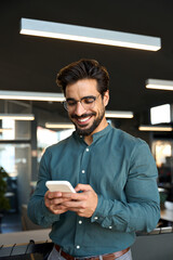 Happy young Latin business man executive, businessman manager standing in office holding smartphone...