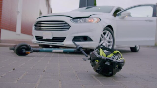 Close-up of a protective head helmet spinning down on the asphalt after a serious road accident from an electric scooter and a luxury white car in a residential area. Concept of road accidents