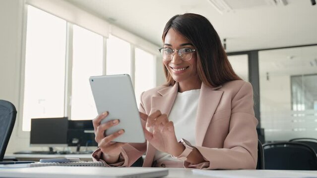 Young happy busy professional African American business woman company manager sales executive wearing suit and glasses, using digital tablet working sitting at desk in office looking at tab.