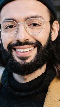 Close-up vertical portrait of positive young hipster man smiling at camera. Handsome trendy bearded guy with hat and glasses standing outdoors. Business people concept