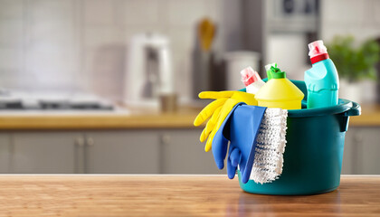 Bucket with cleaning items on wooden table and blurry modern kitchen background. Washing set...