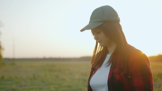 A woman in a cap looks around, walks outdoors at sunset through a green field
