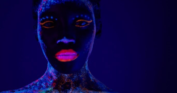 black serious lady with body art glowing in ultraviolet light. beautiful woman's face painted in fluorescent powder,isolated dark purple space. beauty, fashion concept. Beauty industry, cosmetology