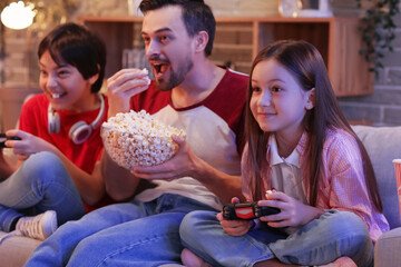 Father with his little children eating popcorn while playing video game at home in evening