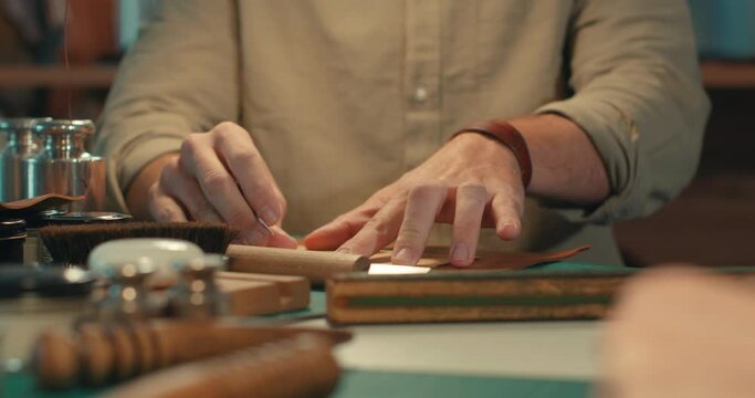Young man placing template on leather piece, putting weight onto pattern, using awl marking outline of leather detail on scale cutting mat indoors. Work tools lying on work table on foreground