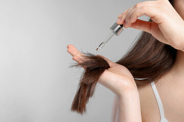 Woman applying essential oil onto hair on light grey background, closeup