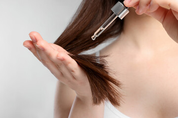 Woman applying essential oil onto hair on light grey background, closeup
