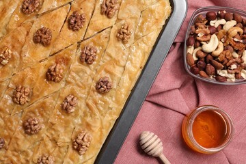 Delicious baklava with walnuts in baking pan, honey and nuts on red cloth, flat lay