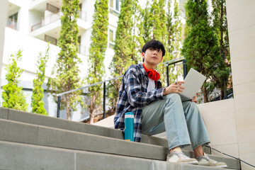 Young Asian man freelancer sitting in public park in the city working business freelance work job on laptop computer. Digital nomad people working anywhere on gadget device with online network.