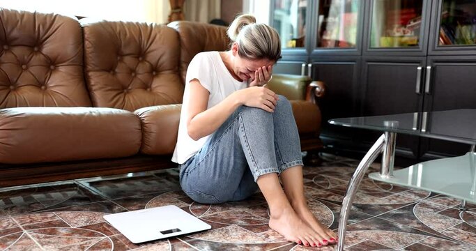 A woman at home, sitting on the floor cries near the scales, a close-up, slowmotion. Weight gain, bad diet