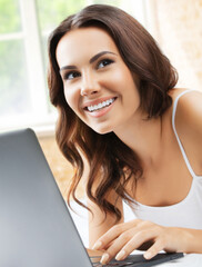 Image of beautiful happy smiling brunette woman using laptop, looking away with smile, at home, bed room, near window.