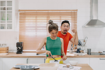 Asian couple. Funny shocked face reaction of husband looking at wife cooking.
