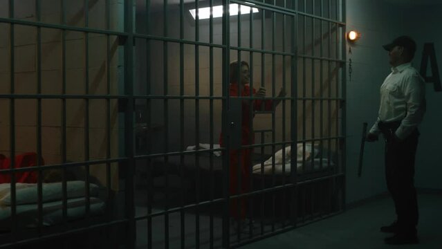 Jailer with police baton stands near prison cell, talks to female prisoner in orange uniform through metal bars. Women serve imprisonment terms in jail. Detention center or correctional facility.