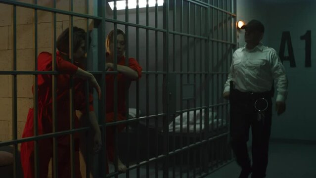 Prison worker walks the corridor. Female prisoners stand behind metal bars in prison cells, give high-five. Women serve imprisonment term for crimes in jail. Detention center or correctional facility.