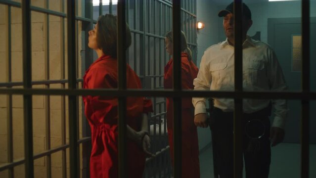 Female prisoners, inmates in orange uniforms stand facing the metal bars in front of prison cells. Jailer walks, talks to women, watches criminals in jail. Detention center or correctional facility.