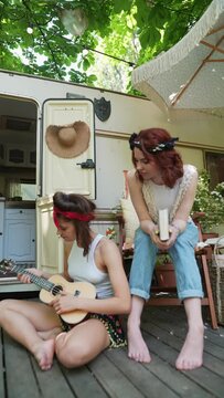 Two hippie girls by the trailer, one playing the guitar, and the other with a book in hand.