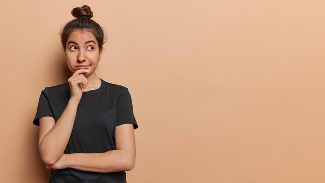 Horizontal shot of lovely pensive Iranian girl keeps hand on chin concentrated asie has thoughtful expression dressed in casual black t shirt isolated over brown background with copy space for promo