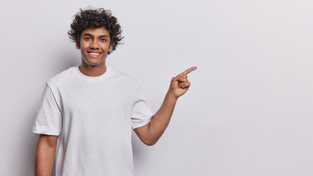 Cheerful Hindu Male Model With Dark Curly Hair Toothy Smile Pointing At Copy Space For Advertisement Or Text Showing Great Deal Or Offer Dressed In Casual T Shirt Isolated Over White Background.