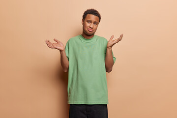 People emotions concept. Studio photo of young confused African male not knowing how to solve difficult problem or task wearing casual clothes standing isolated in centre on beige background