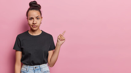 Serious displeased Latin woman with hair bun raises eyebrows looks attentively at camera purses lips has dimple on cheek dressed in casual black t shirt and jeans isolated over pink background - obrazy, fototapety, plakaty