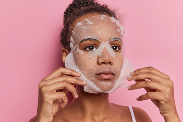 Indoor shot of good looking African girl with dark hair gathered in bun applies moisturising sheet...