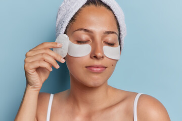 Close up shot of young woman with healthy glowing skin applies beauty patches under eyes and uses face massager undergoes cosmetology procedures wears bath towel on head isolated on blue wall