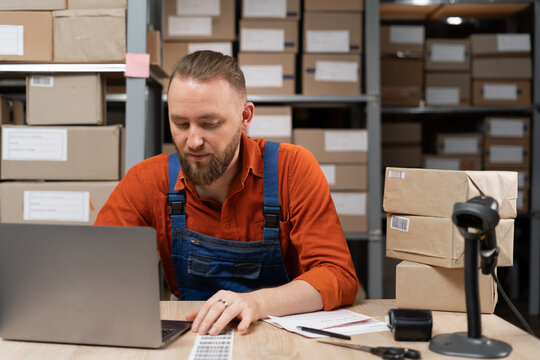 Manager using laptop computer to check inventory while working in warehouse retail center with cardboard boxes. E-Commerce