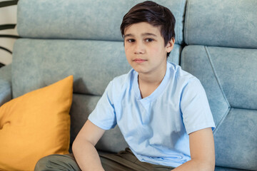 Close-up portrait of a teenage boy wearing a blue t-shirt. sitting on the couch in the room, looking at the camera