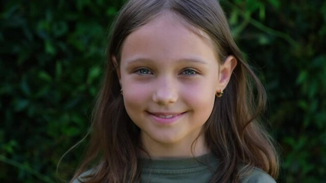 Portrait of a beautiful brunette little teenager girl looking at the camera and smiling, standing on the street against the backdrop of greenery on a summer day. Young curious child raises his eyes