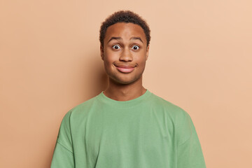 Portrait of surprised dark skinned man reacts to something shocking wears casual green tshirt has widely opened eyes poses against brown background. Human reactions and face expresssions concept